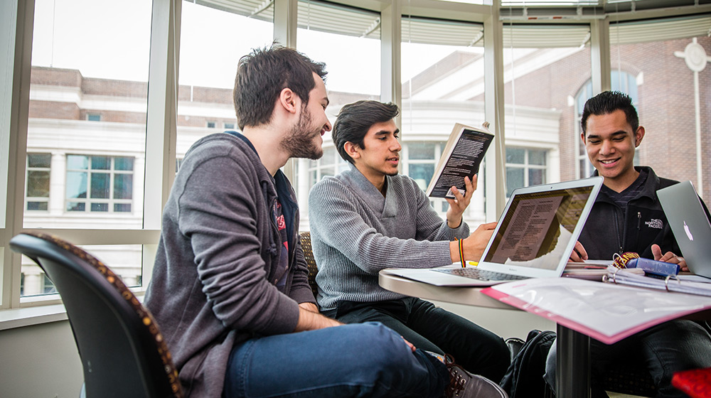 Three young men sit at a round table in a sunny room with large windows. They are engaged in a discussion, with laptops and books in front of them. One man is pointing to a page in a book, while the others listen attentively.